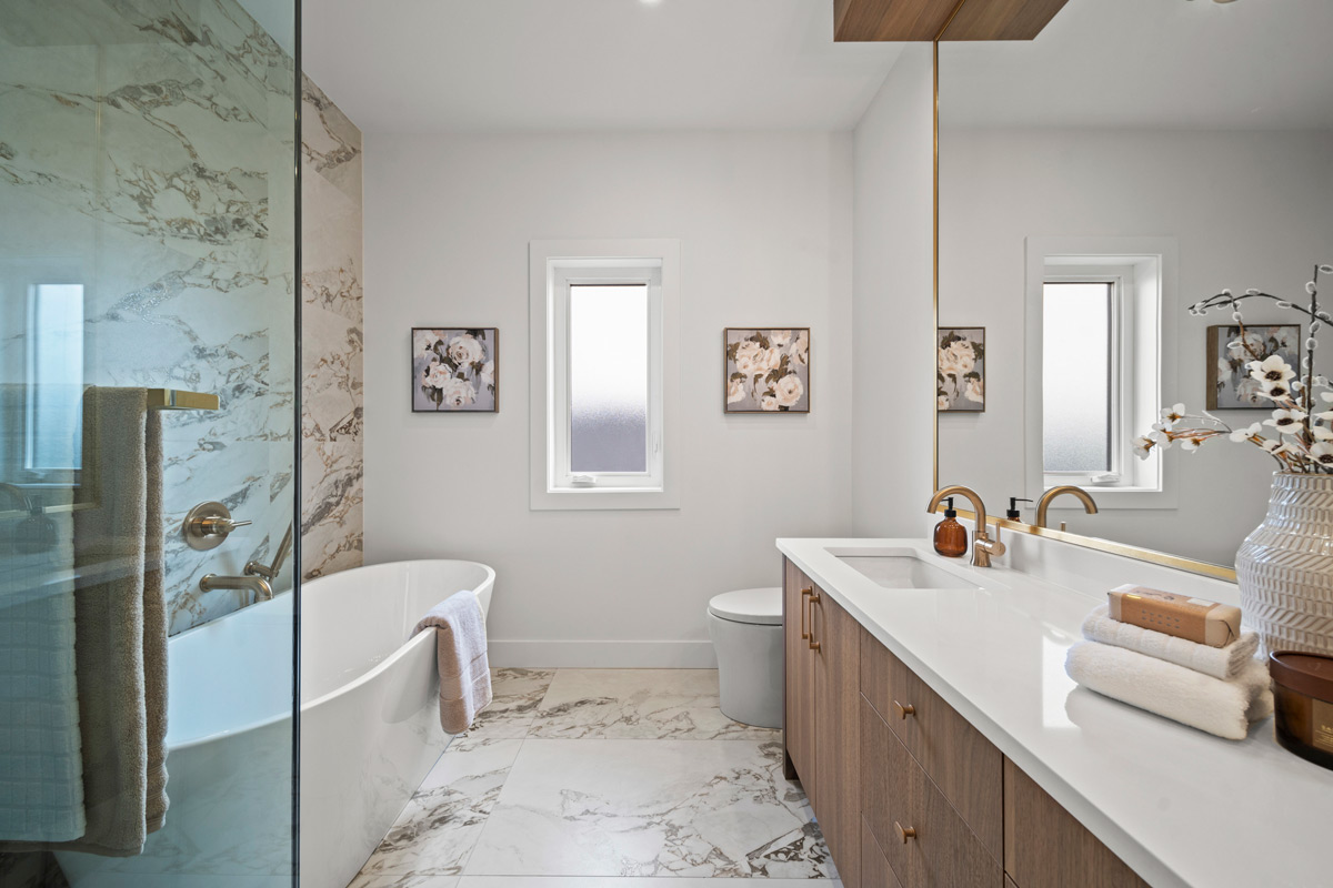 Built by Asthan Homes in Burnaby, BC, modern bathroom with a freestanding white bathtub, marble tile flooring and wall, wooden vanity with white countertop, large mirror, and decorative floral artwork.