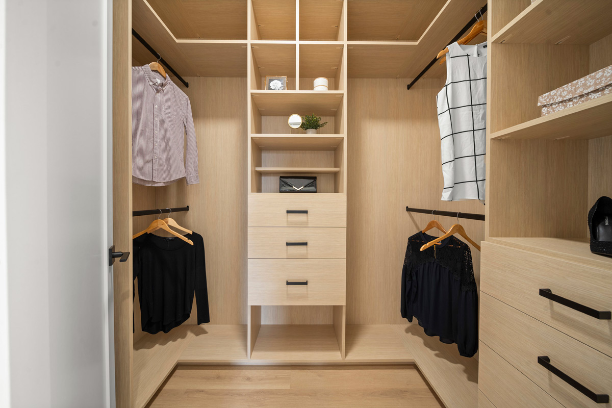 Built by Asthan Homes in Burnaby, BC, walk-in closet with light wood shelving, drawers, and hanging clothes including shirts and dresses.