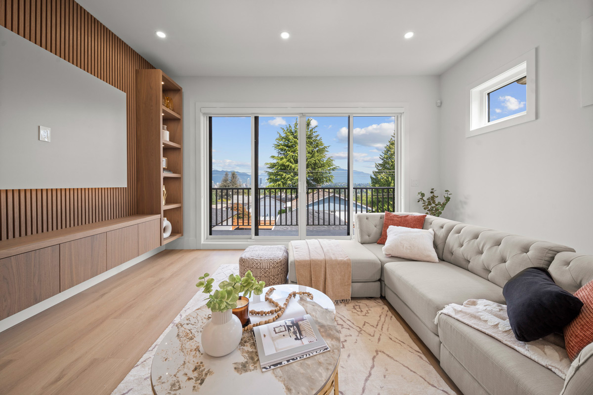Built by Asthan Homes, bright modern living room with light gray tufted sectional sofa, decorative pillows, round marble coffee table, wood shelving, and large sliding glass doors with a tree and mountain view.