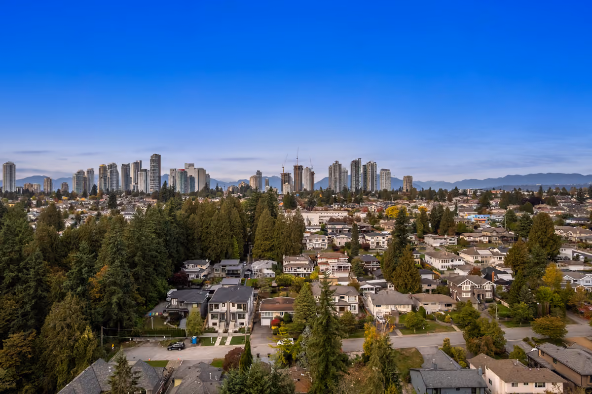 Aerial view of a suburban neighborhood with houses, tall evergreen trees, and a distant city skyline of Burnaby, BC under a clear blue sky.