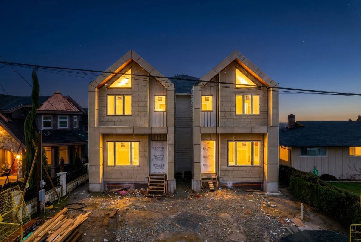 Two-story duplex under construction at dusk with illuminated windows and bare ground in front.