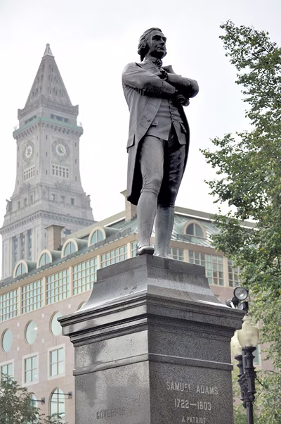 Pedestal-mounted bronze statue of historical figure with cloudy sky and clock tower behind.