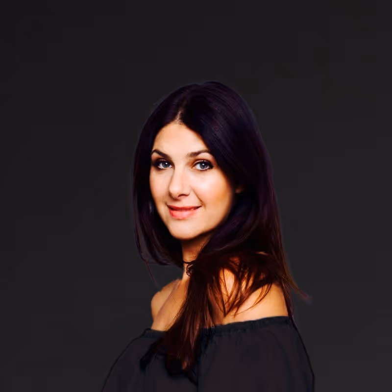 Portrait of a smiling woman with long dark hair wearing an off-the-shoulder black top against a dark background.