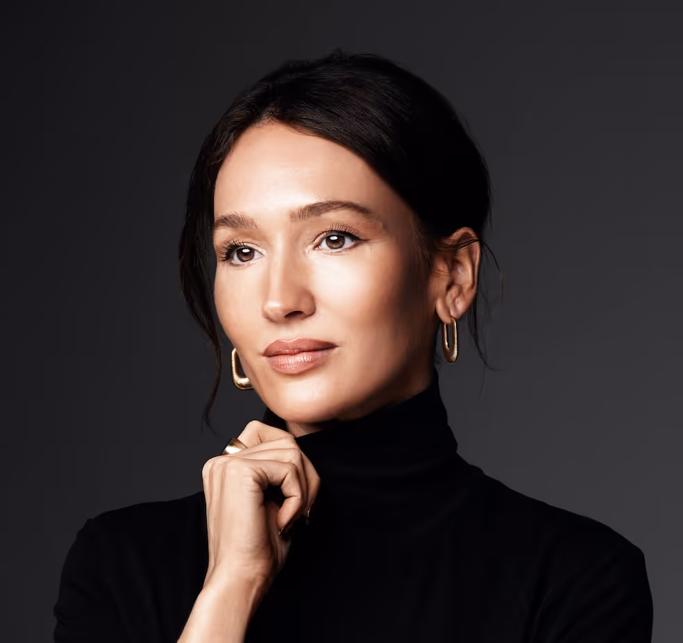 Woman with dark hair wearing gold hoop earrings and a black turtleneck, resting her chin on her hand against a dark background.