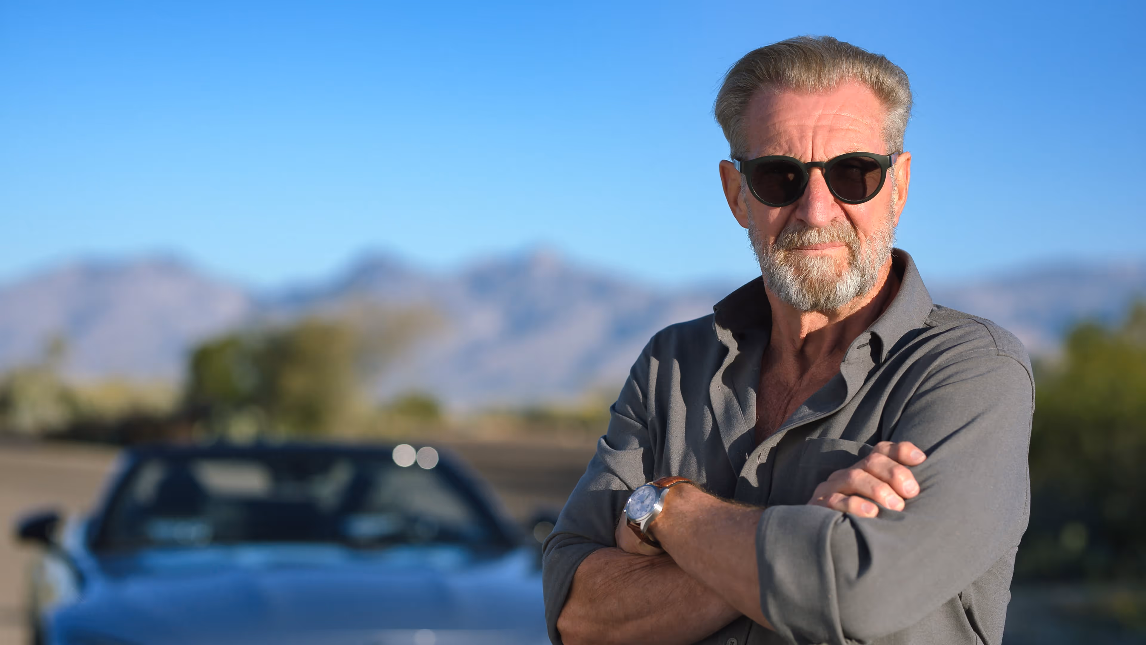 Black and white photo of an older man with a beard and sunglasses driving a car, resting his hand on the steering wheel.