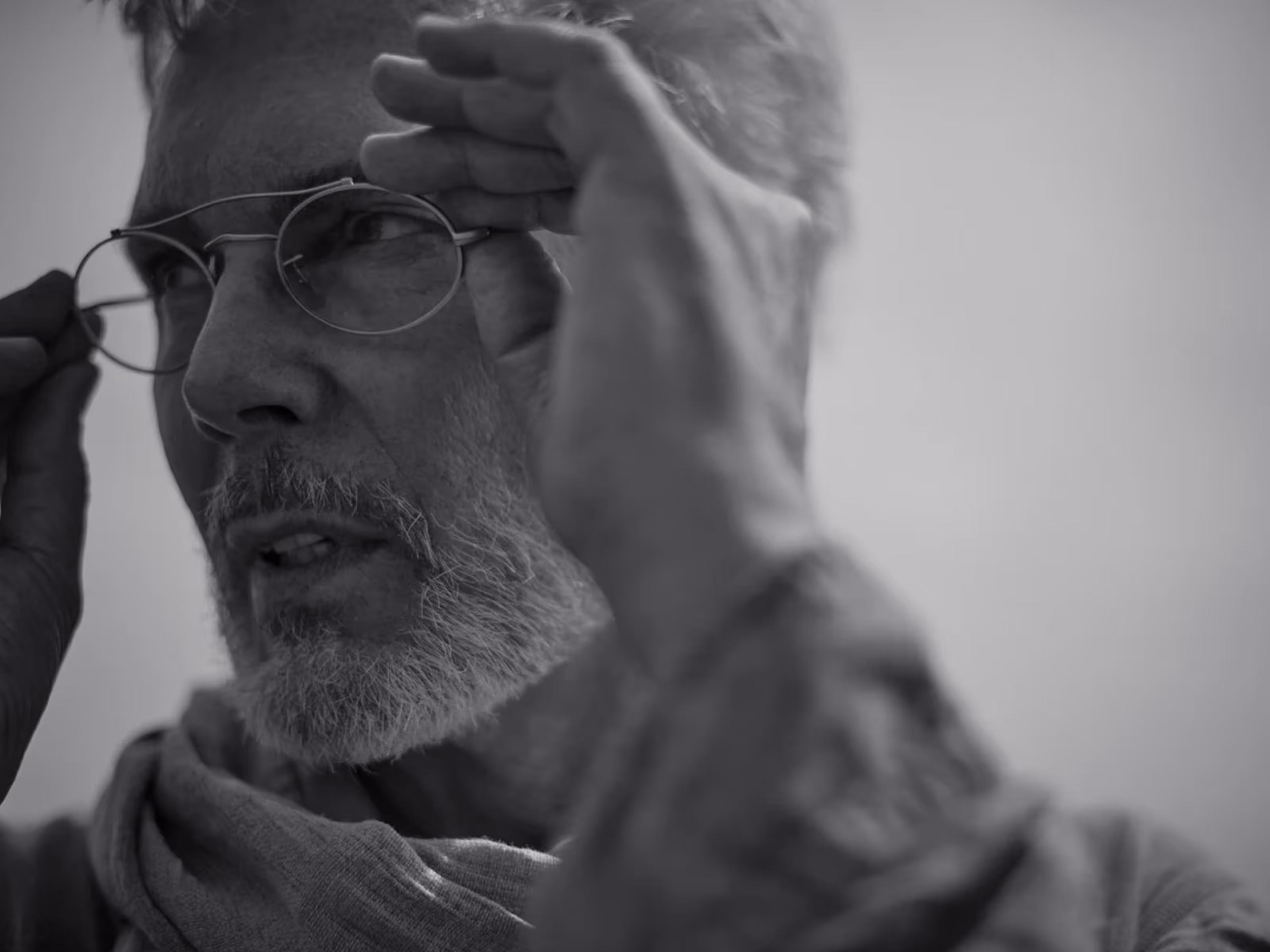 Close-up black-and-white photo of an older man with a beard adjusting round glasses.