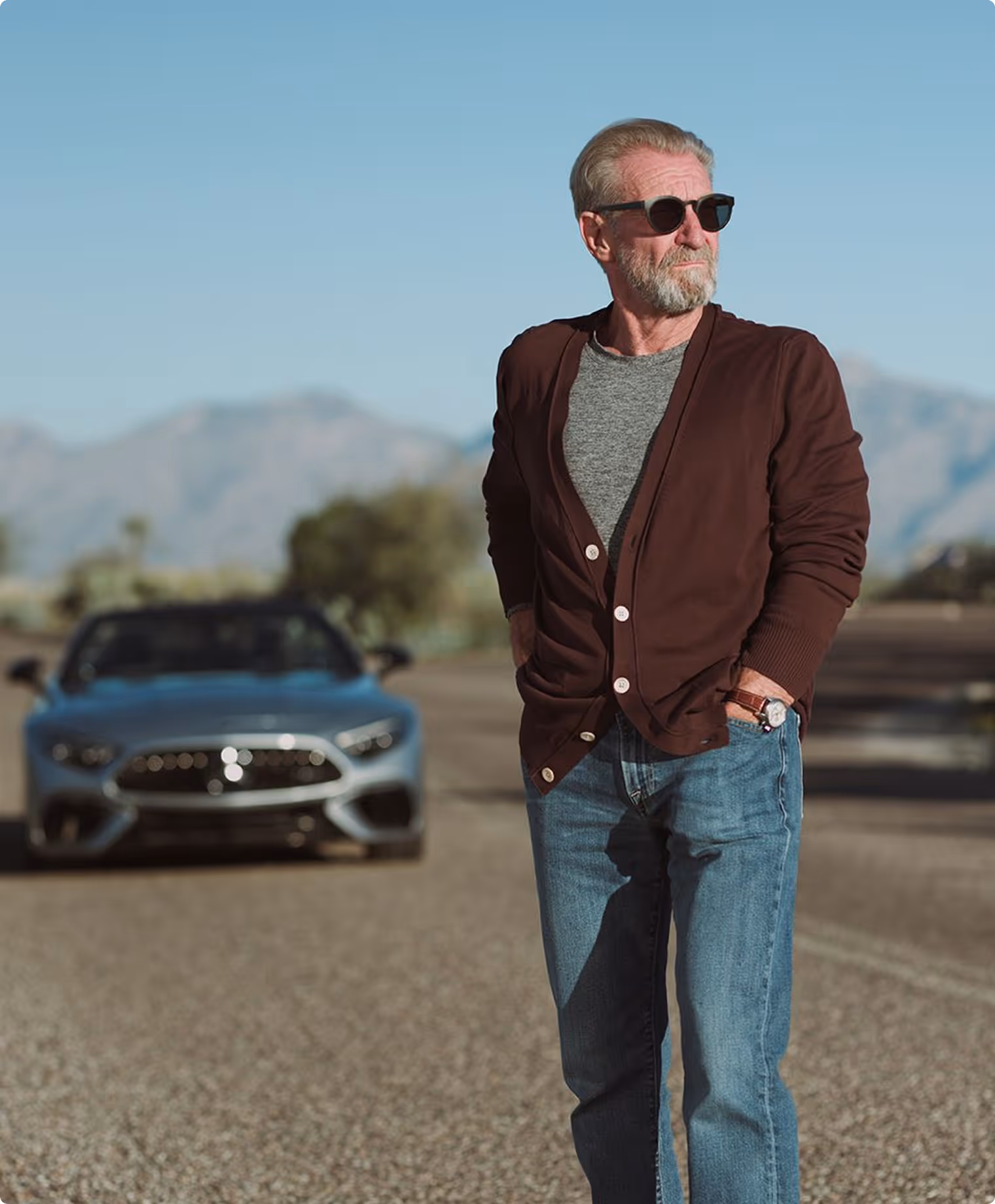 Older man with gray hair and beard wearing sunglasses, a brown cardigan, and jeans standing on a paved road with a blurred sports car and mountains in the background.