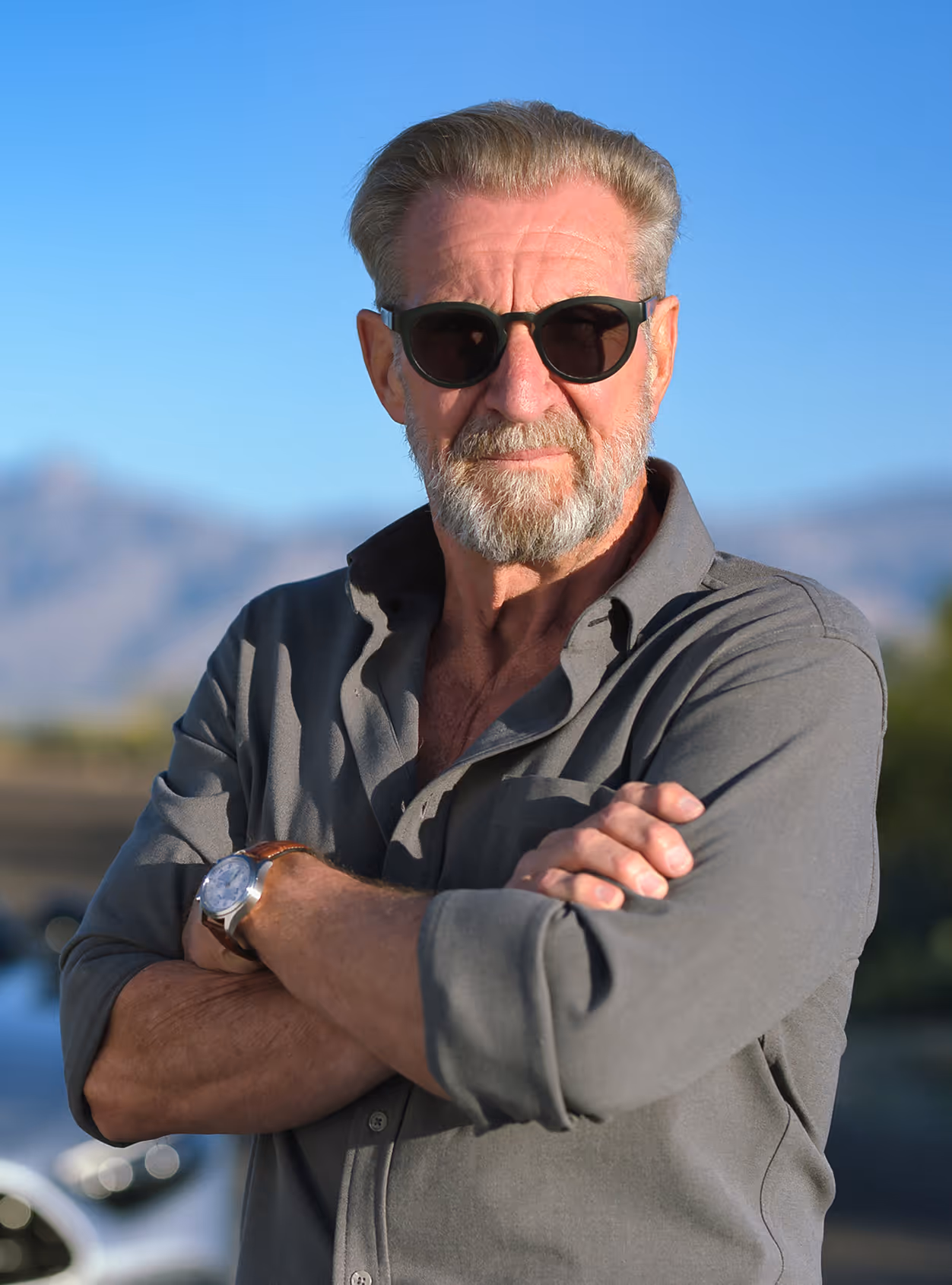 Older man with gray beard and sunglasses, wearing a gray button-up shirt with sleeves rolled, standing outdoors with arms crossed.