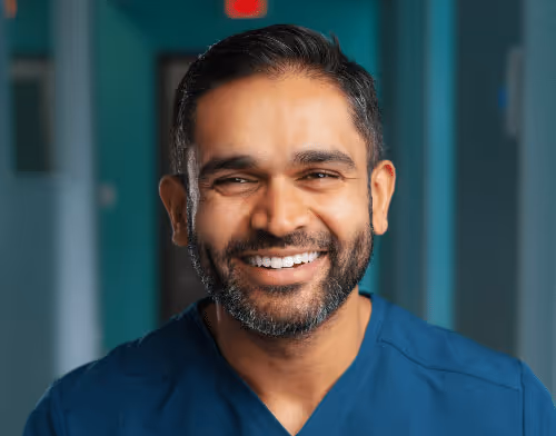 Smiling male healthcare professional with beard and dark hair wearing blue scrubs in a clinical setting.