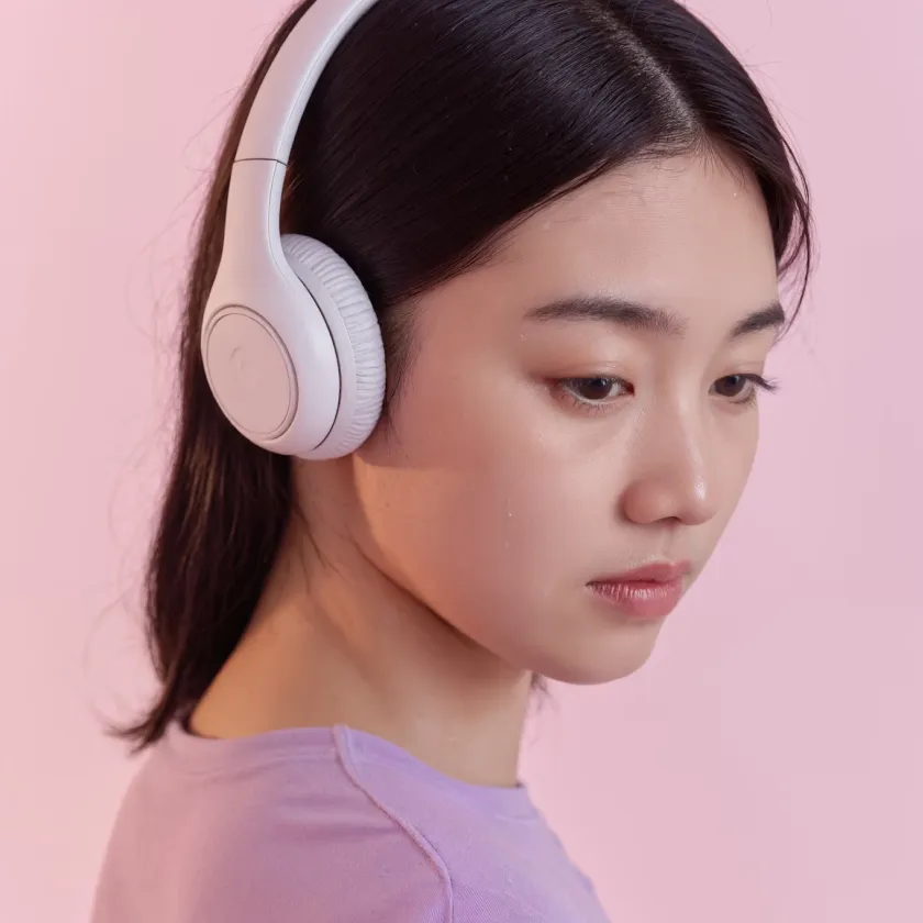 Young woman with dark hair wearing white headphones and a lavender top, looking downward against a pink background.