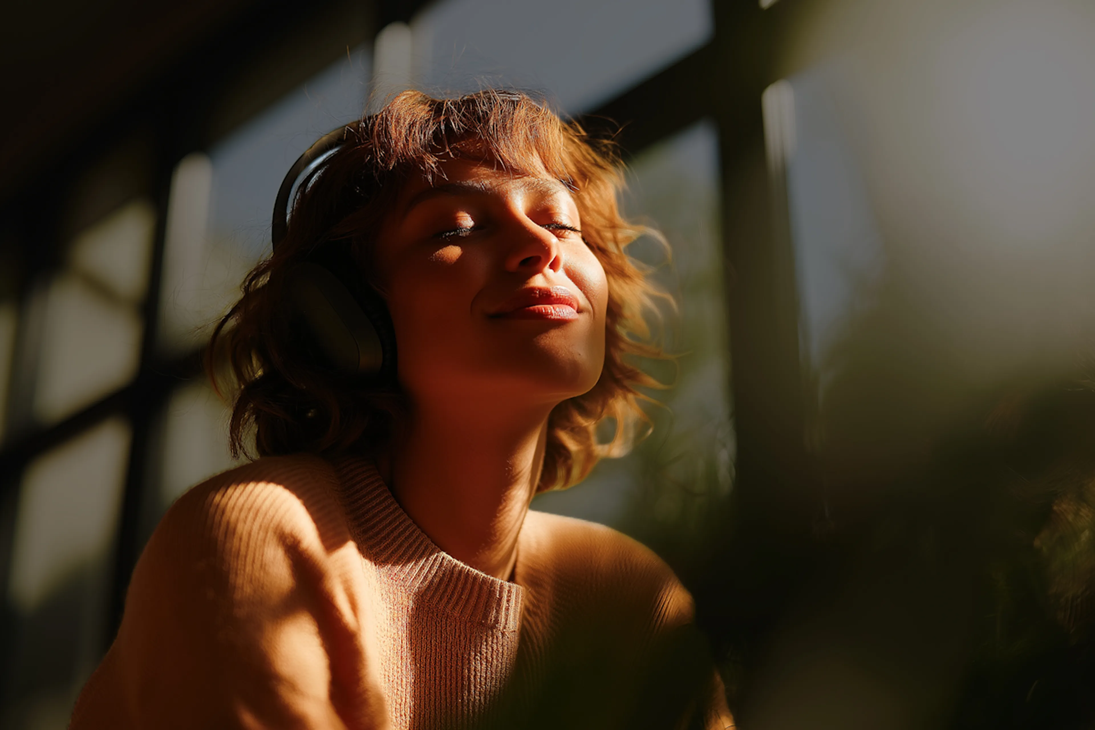 Woman with short curly hair wearing headphones and a peach sweater, enjoying music with eyes closed in warm sunlight.