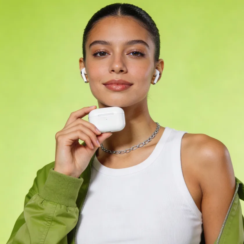 Young woman wearing wireless earbuds and holding the earbuds' charging case, posing against a light green background.