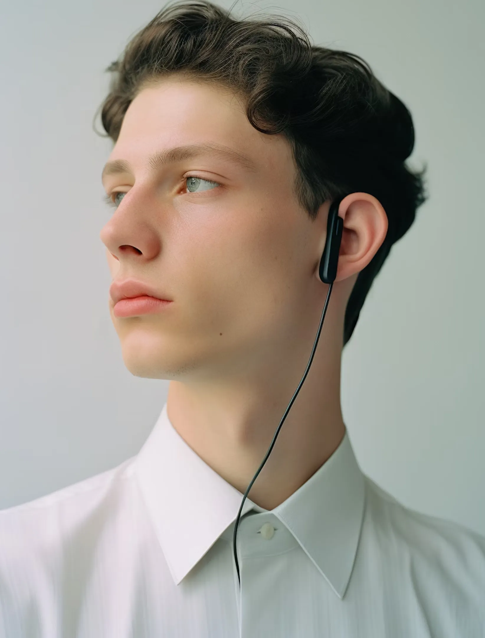 Young man in a white collared shirt wearing black wired earphones, looking to the side.