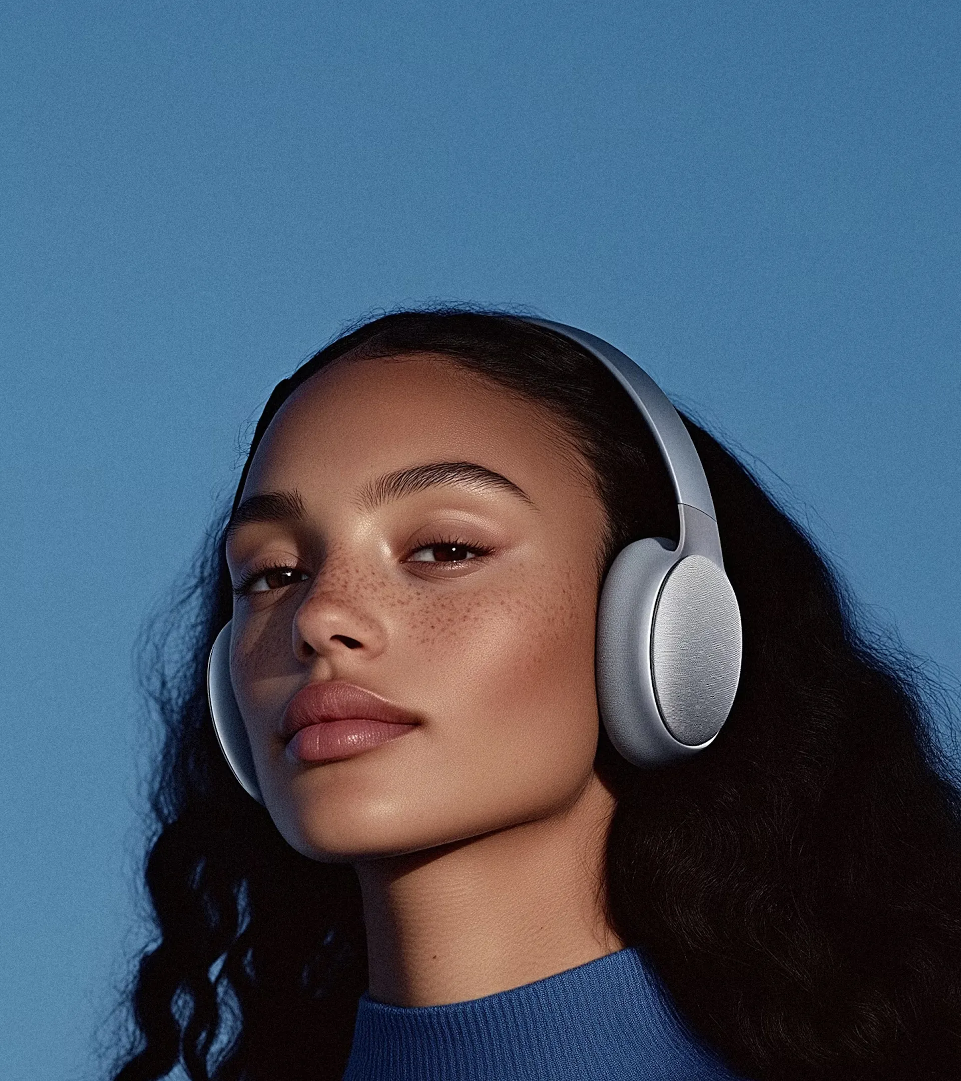 Close-up of a woman with freckles and curly hair wearing silver over-ear headphones against a blue background.