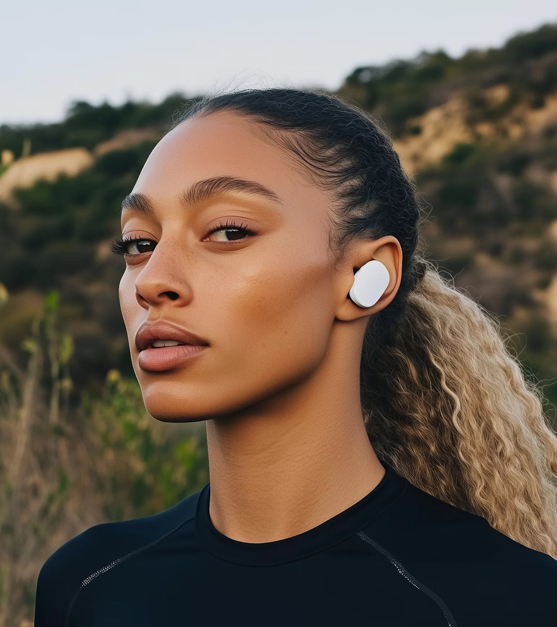 Young woman outdoors wearing a black top and white wireless earbuds with a natural background.