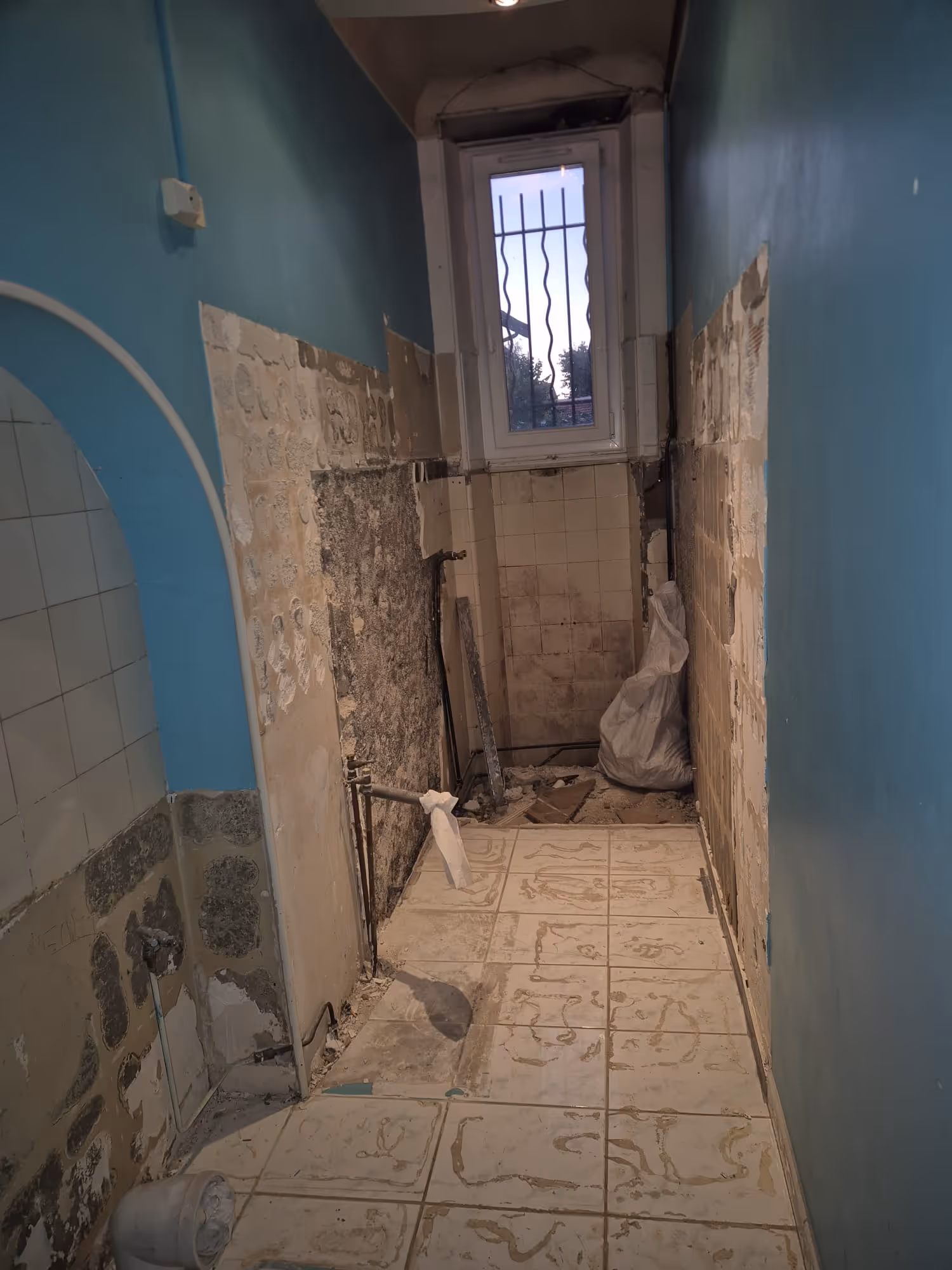 Sol en carrelage bleu et blanc avec restes de travaux de plomberie dans une pièce aux murs partiellement carrelés et endommagés.