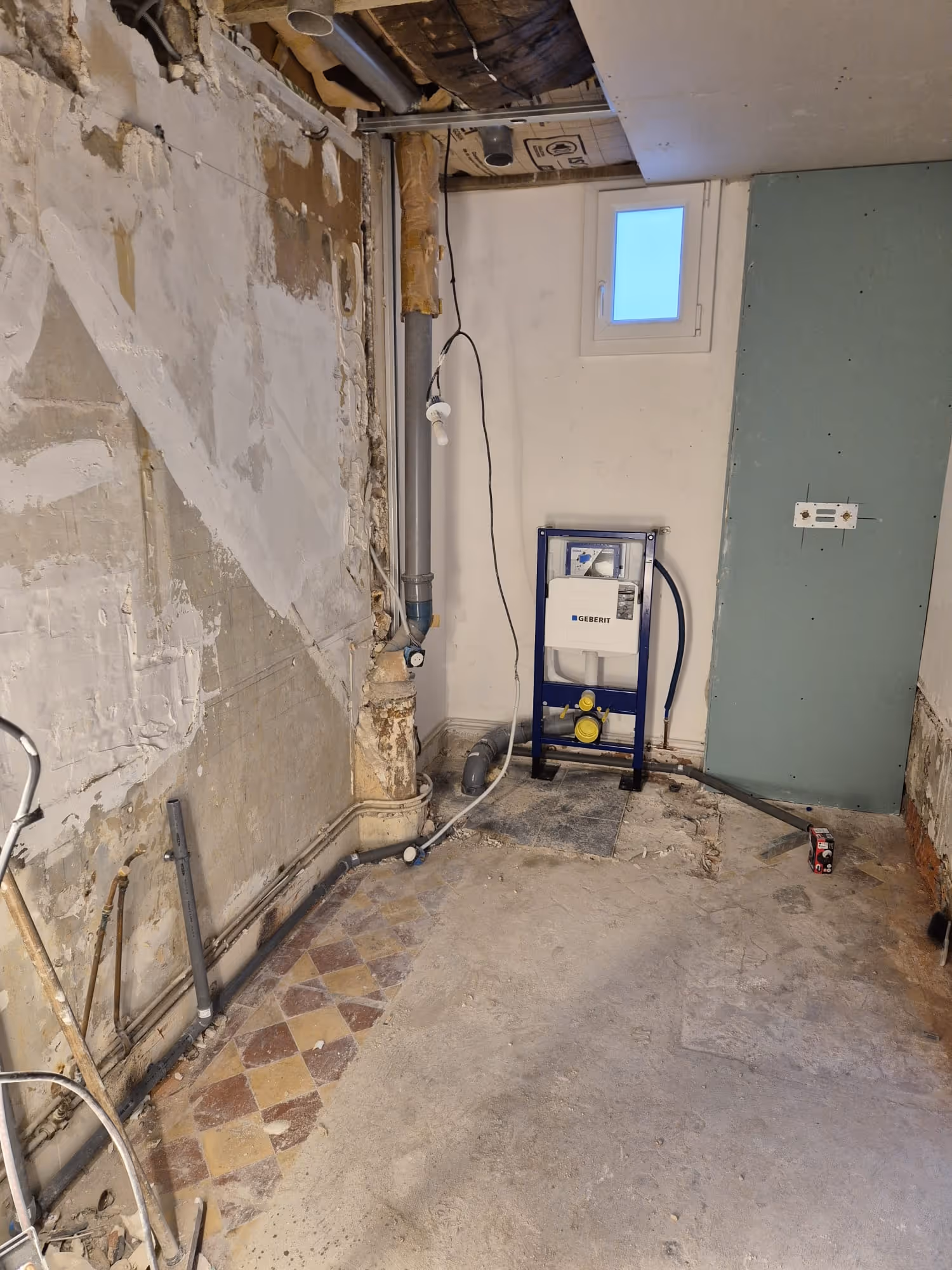 Pièce en rénovation avec murs partiellement dégarnis, tuyauterie apparente et montage d'une structure de chasse d'eau encastrée près d'une petite fenêtre.