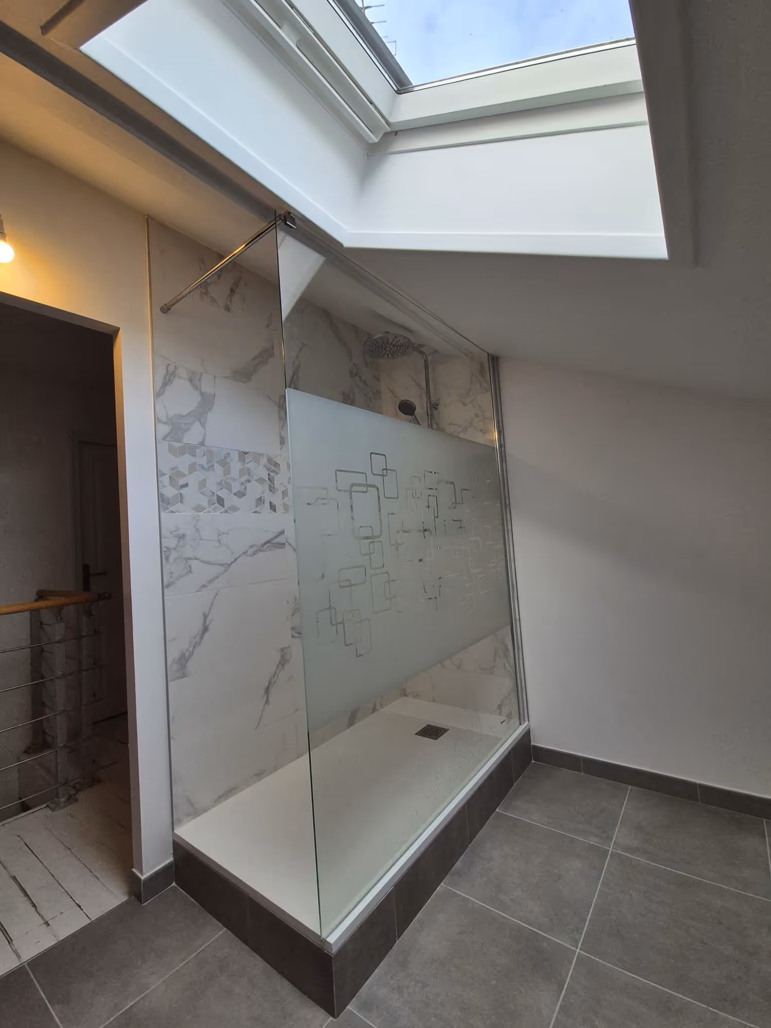 Douche moderne avec une paroi vitrée gravée de motifs géométriques sous une fenêtre de toit.