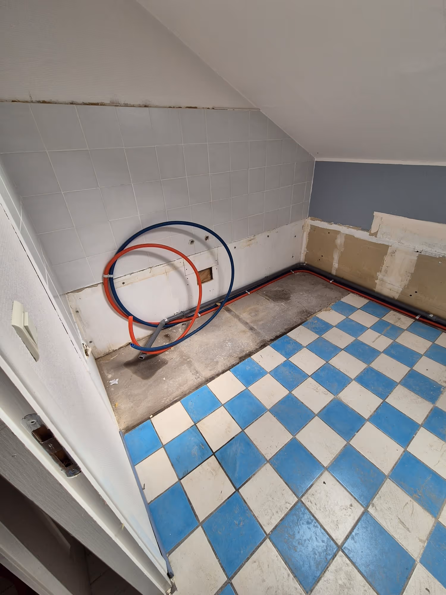 Sol en carrelage bleu et blanc avec restes de travaux de plomberie dans une pièce aux murs partiellement carrelés et endommagés.