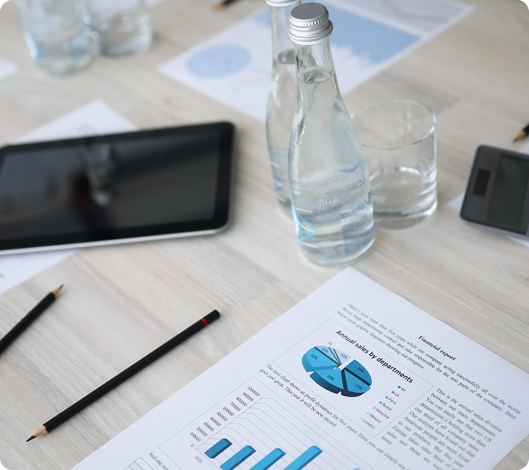 Office meeting table with documents showing charts, a tablet, a pencil, and glass bottles of water arranged on a desk.