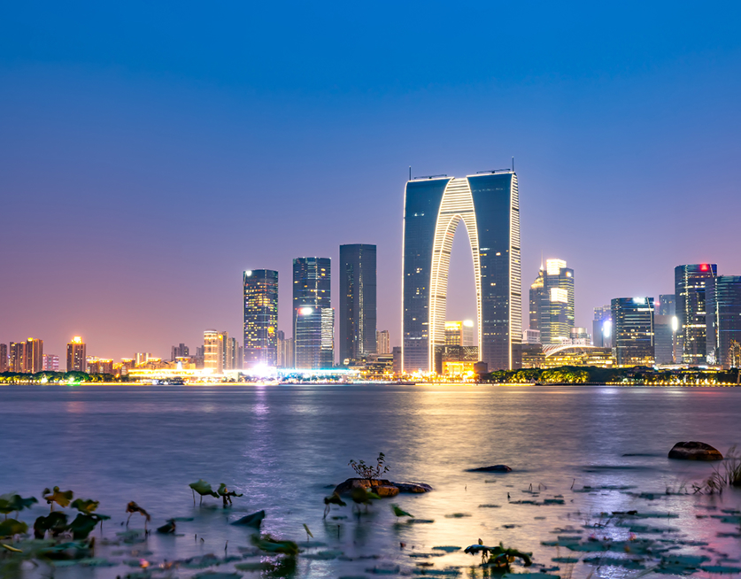 Panoramic view of a modern city skyline at dusk, with illuminated skyscrapers reflected on the water in the foreground.