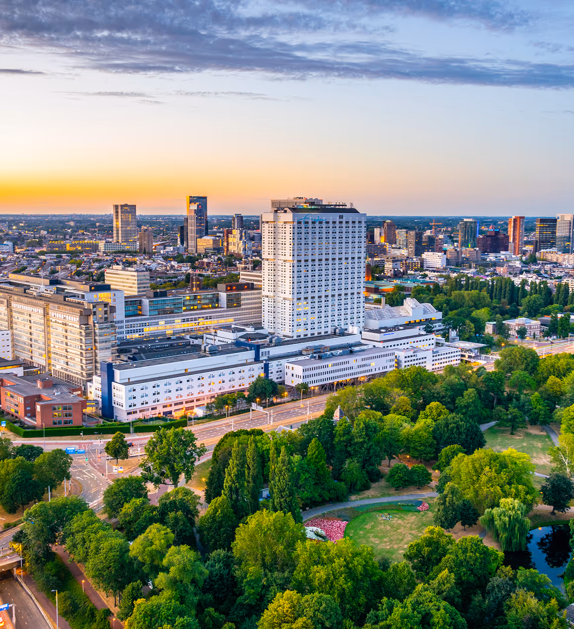 Aerial view of a cityscape at sunset featuring a large white hospital building surrounded by trees and roads.