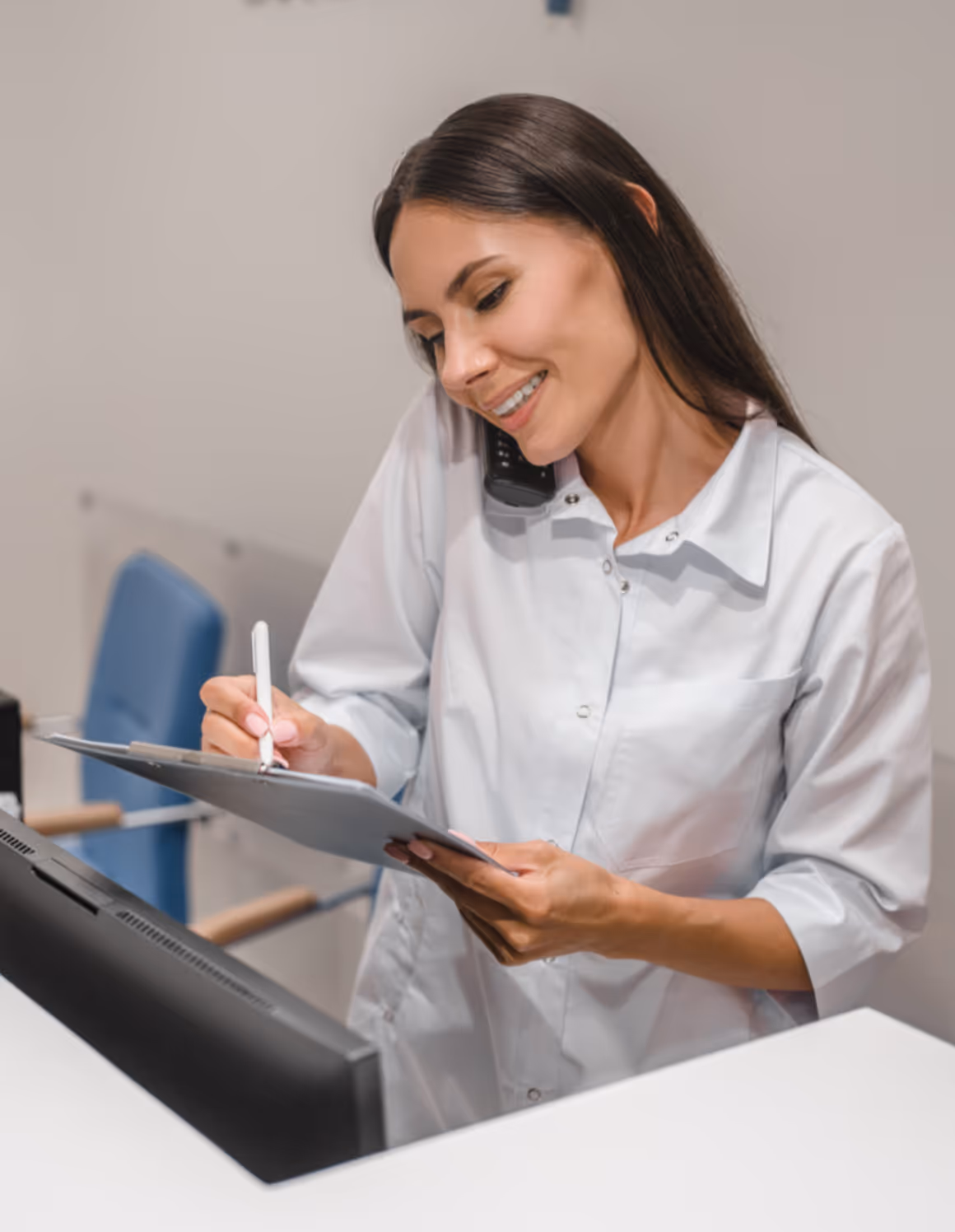Smiling female healthcare professional in white coat talking on phone and writing on a clipboard at a reception desk.