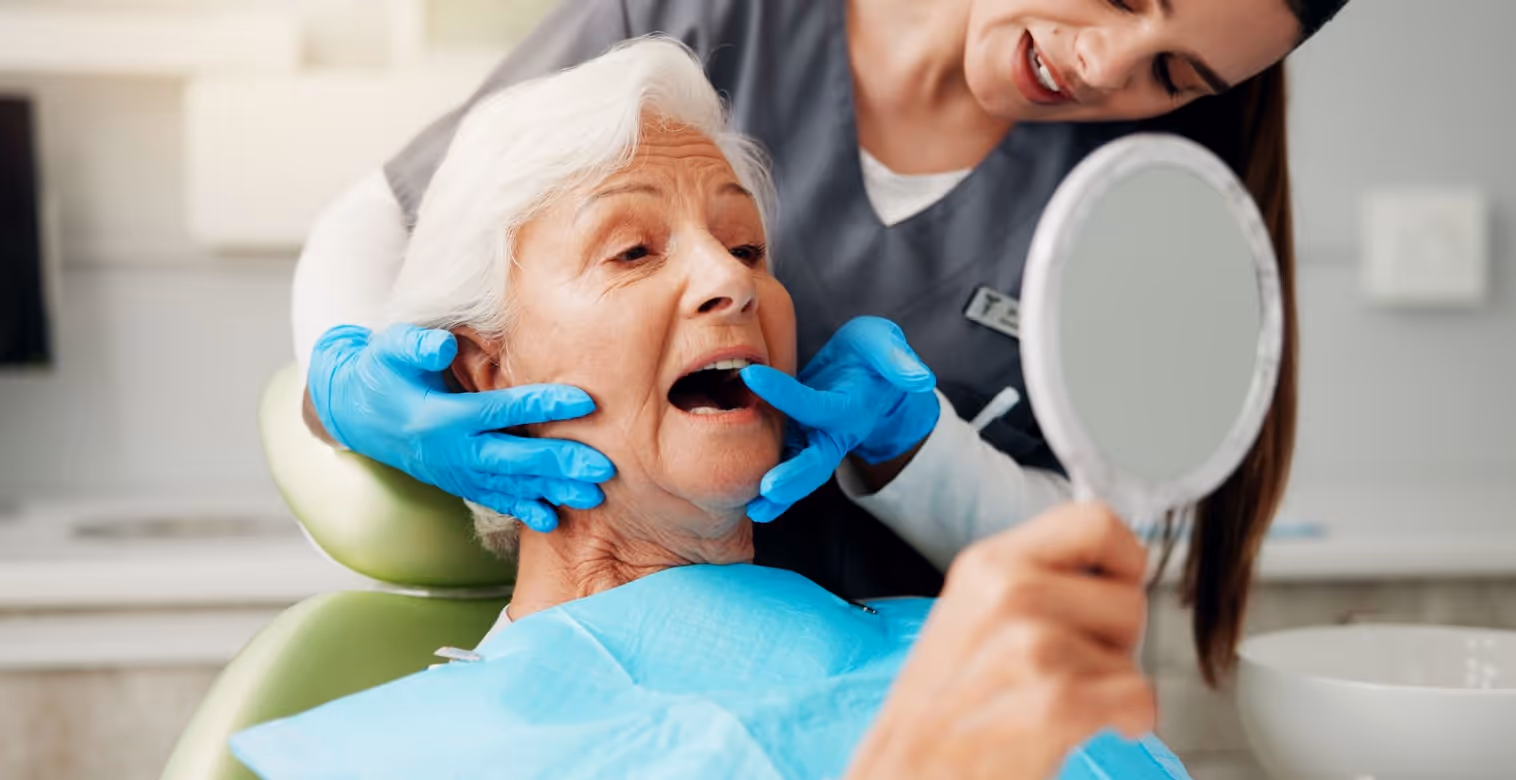 Dental assistant helping an older woman examine her mouth using a handheld mirror