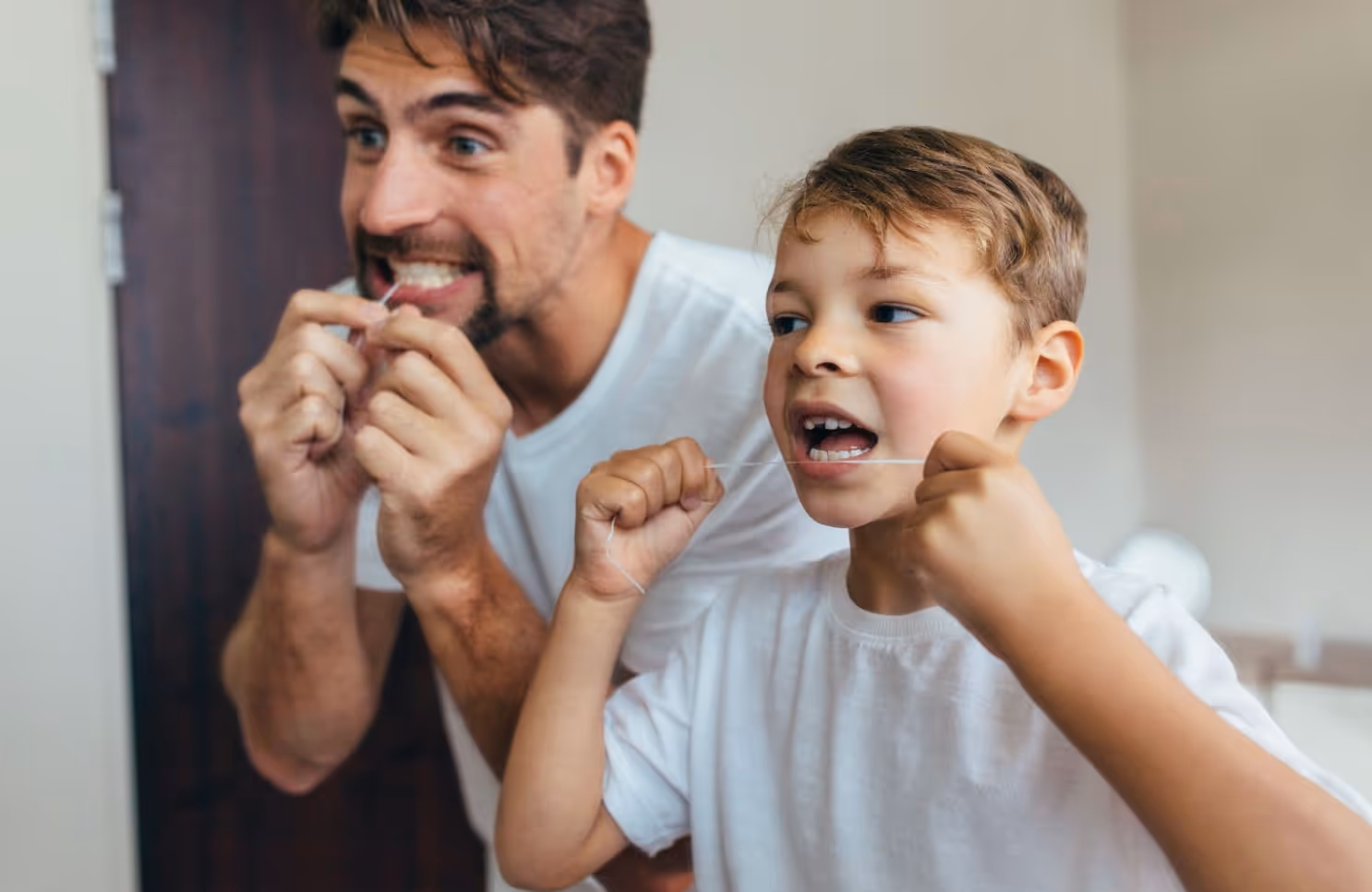 Father and young son flossing their teeth together in front of a bathroom mirror
