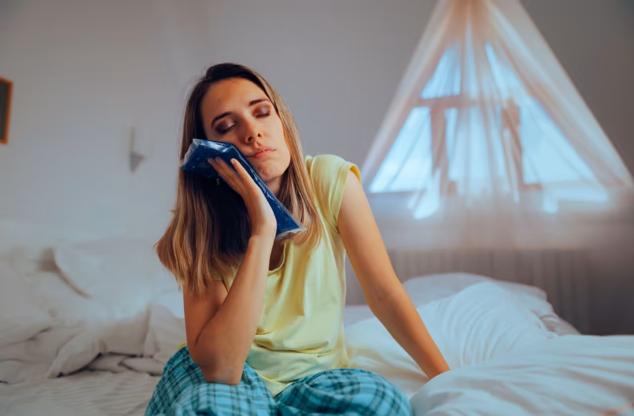 Woman sitting in bed holding an ice pack to her cheek to ease dental discomfort