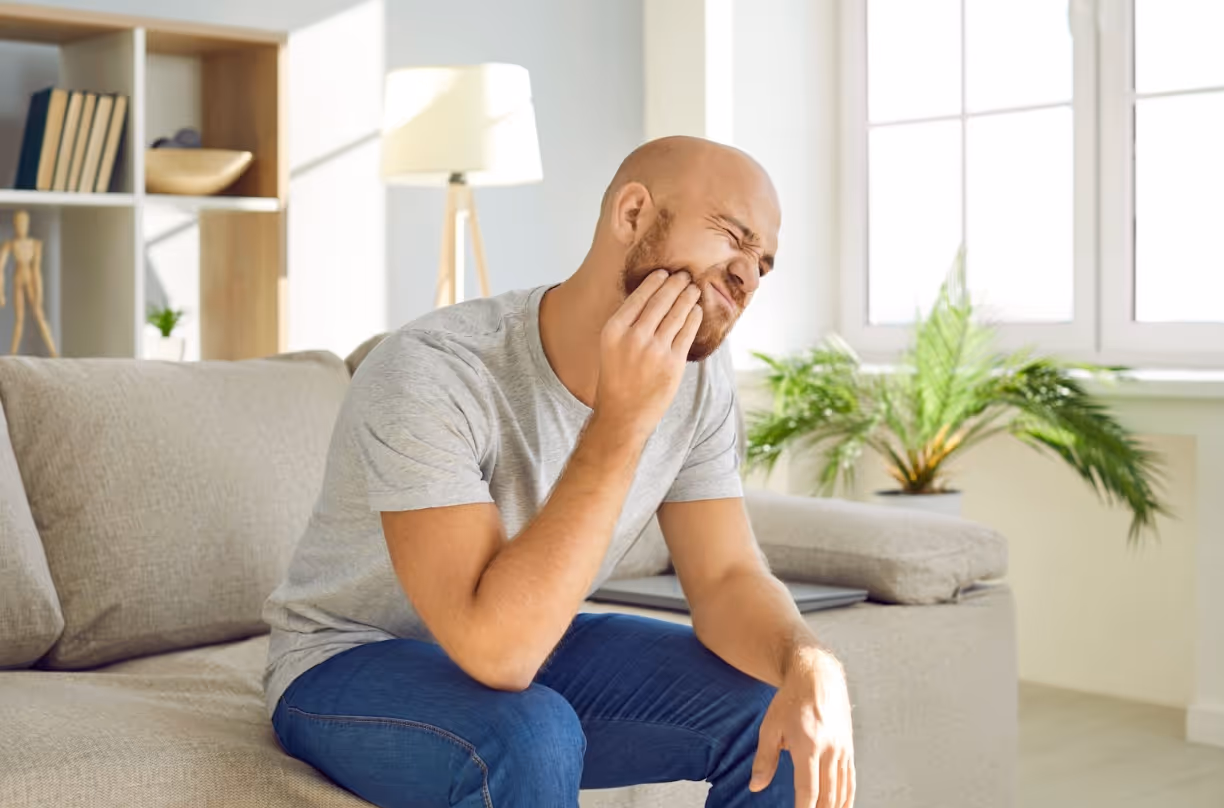 Man sitting on a couch holding his jaw due to tooth pain.