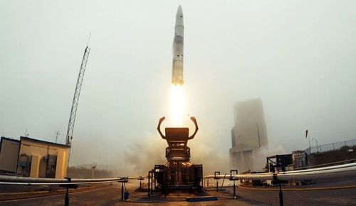 Rocket launching from a ground platform with smoke and flames beneath it and industrial buildings in the background.