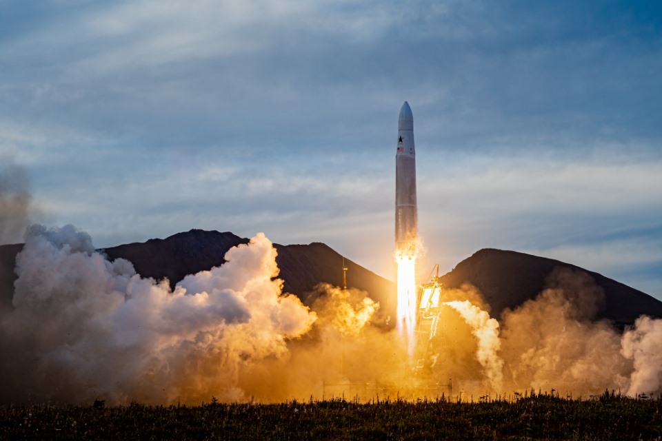 A rocket launching with flames and smoke billowing at the base, mountains in the background under a cloudy sky.