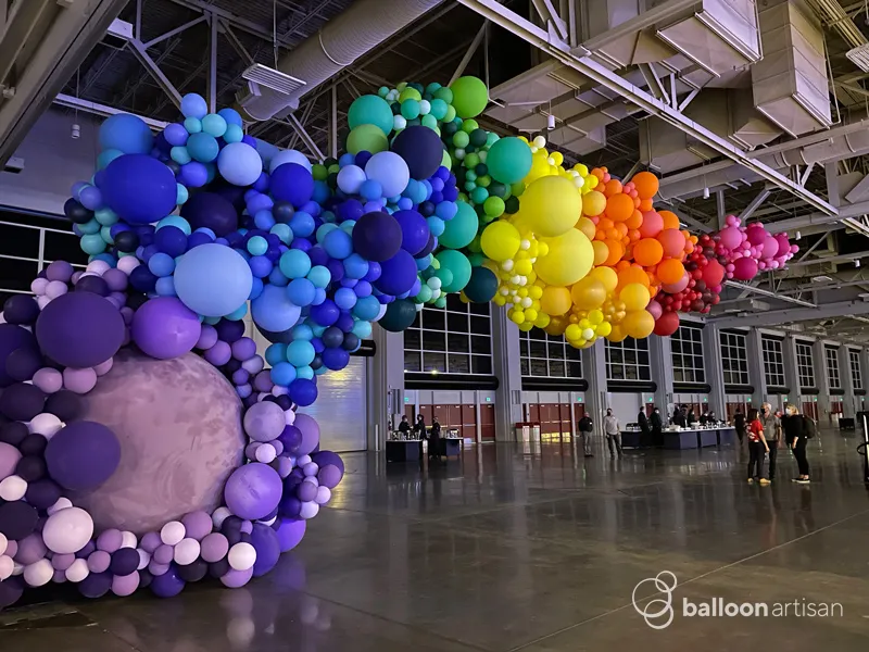 A large rainbow balloon arch decor installation decorates the entrance of a large convention center for a company party balloon photo backdrop.