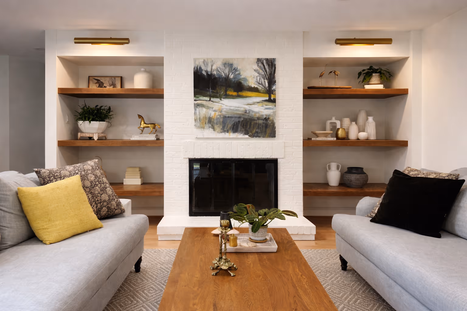 Cozy living room with white brick fireplace, built-in wood shelving, landscape artwork, gray sofas, and wooden coffee table with decorative accents.