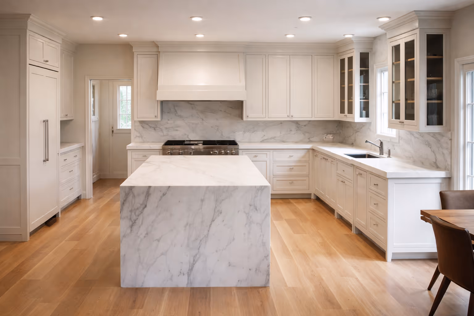 Spacious white kitchen with large marble waterfall island, custom cabinetry, marble backsplash, stainless steel range, and light hardwood floors.