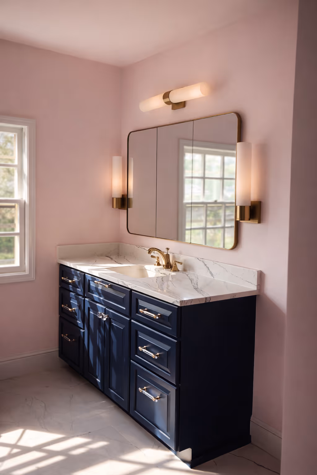 Elegant bathroom vanity with navy blue cabinets, marble countertop, brass faucet, pink walls, and gold-framed mirror with wall sconces.