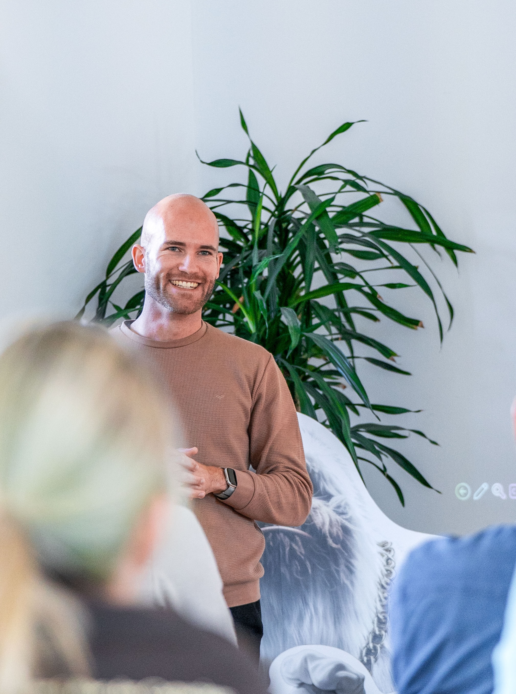 Mann steht vor einer Gruppe und hält eine Präsentation, lächelt in die Kamera; im Hintergrund eine große grüne Zimmerpflanze und eine helle Wand.
