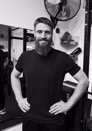 Bearded man in black t-shirt standing with hands on hips in a gym setting, smiling at the camera.