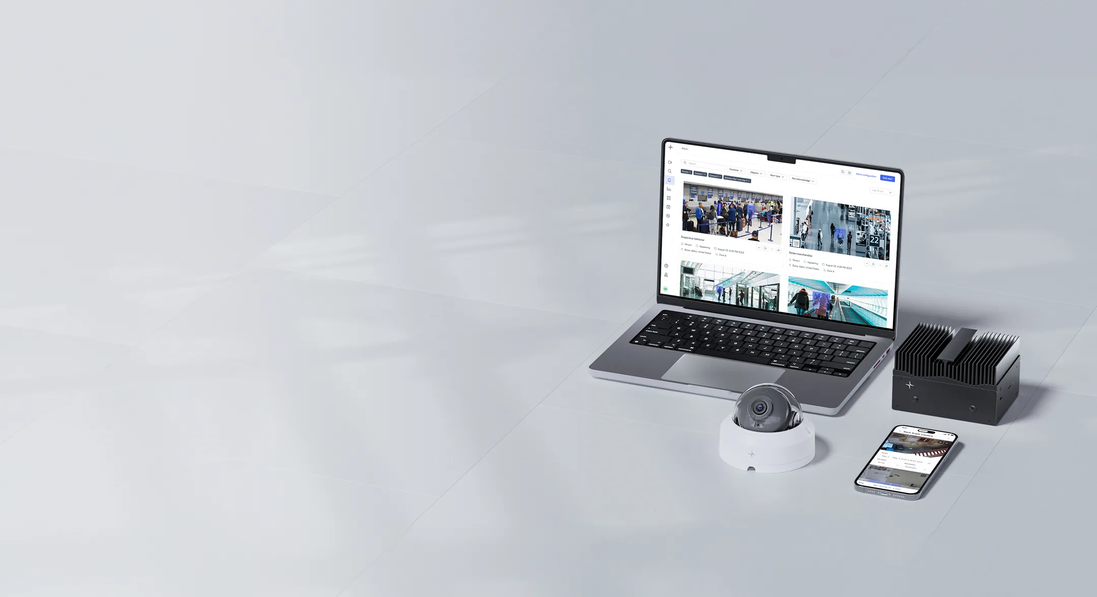 Laptop, security camera, black device, and smartphone displaying surveillance footage on a light-colored surface.