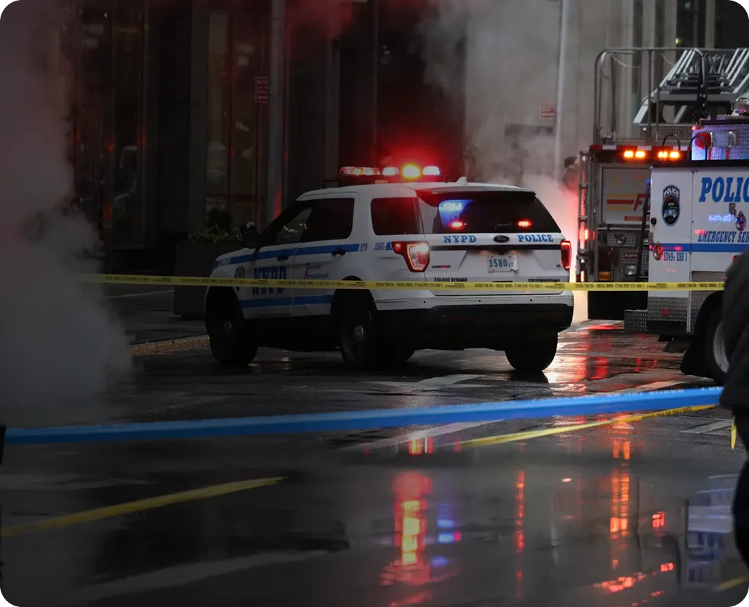 NYPD police vehicle with flashing lights behind yellow caution tape on a wet city street at night with smoke in the background.