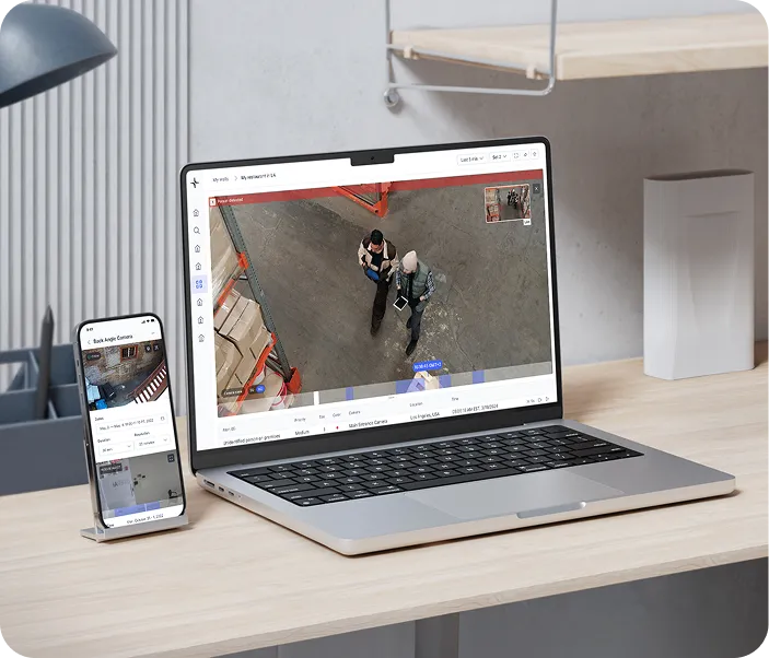 Laptop and smartphone on a desk displaying surveillance camera footage of two people walking in a warehouse.
