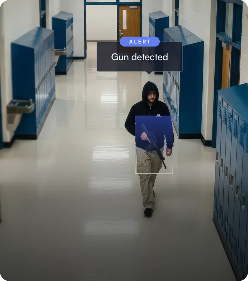 Man in a black hoodie carrying a gun walking down a school hallway with lockers, with an alert message saying 'Gun detected.'