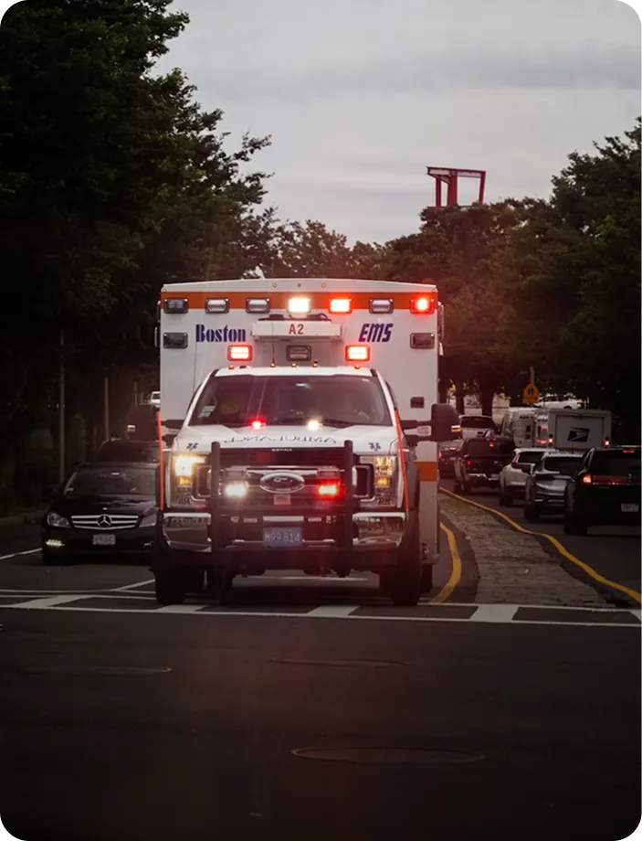 Boston EMS ambulance vehicle with flashing lights driving on city street during dusk.