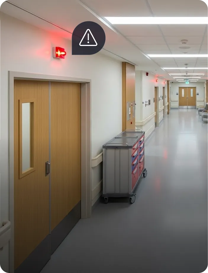 Empty hospital hallway with wooden double doors and a medical cart, emergency warning light glowing red above the doors.