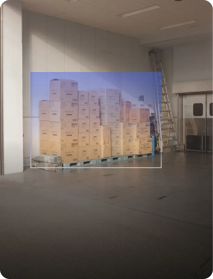 Stacked cardboard boxes on pallets inside a warehouse with a metal ladder and sliding doors in the background.