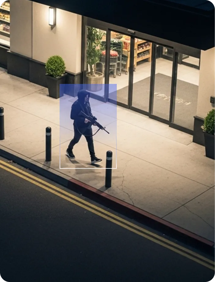 Person wearing a hoodie and mask carrying a rifle walking on a sidewalk near a building entrance at night.
