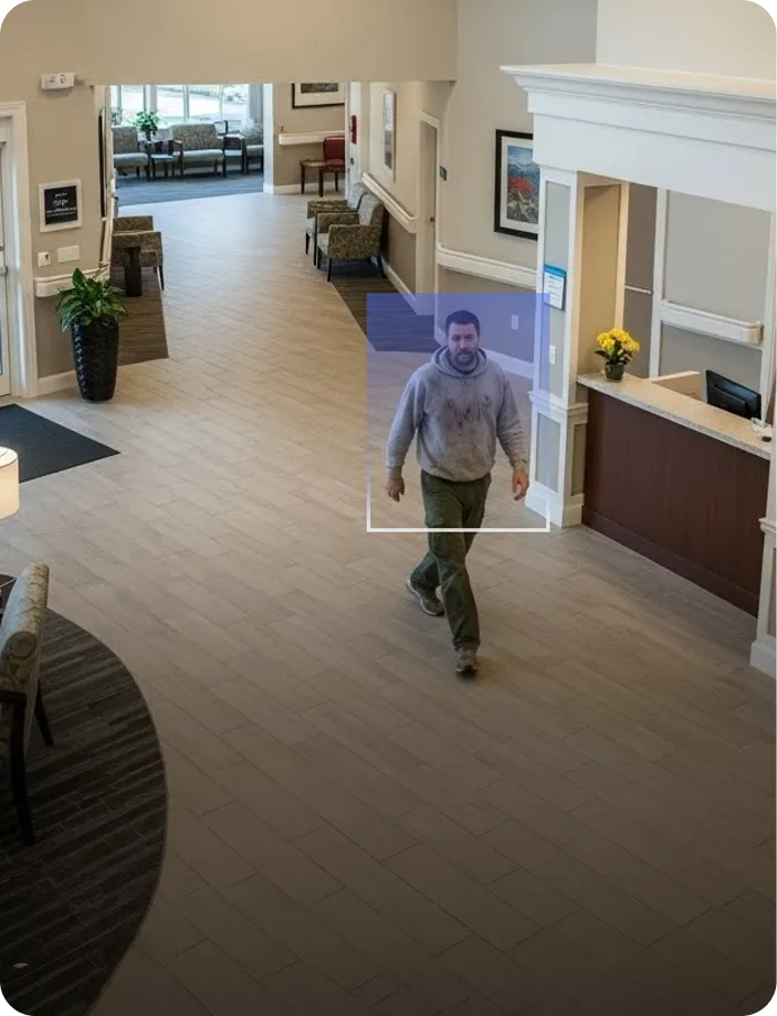 Man in a gray hoodie and green pants walking through a spacious waiting area with chairs, plants, and a reception desk with yellow flowers.