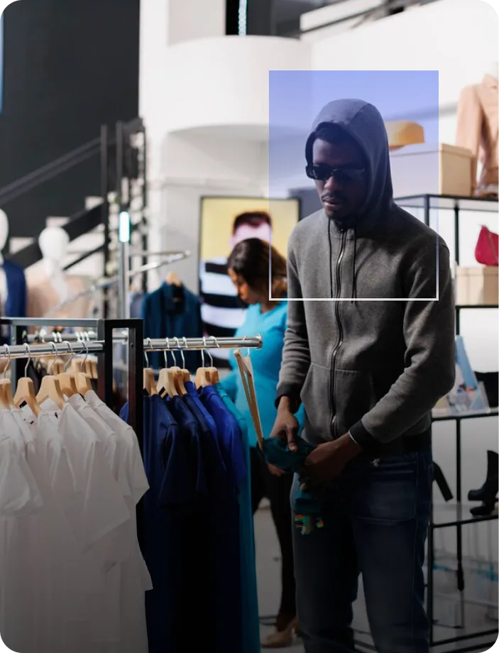 Man in a hoodie and sunglasses browsing clothing rack with shirts inside a store, with a woman shopping in the background.