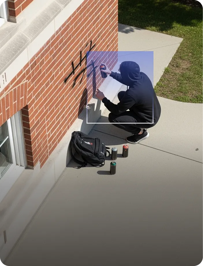 Person wearing a black hoodie crouched on a sidewalk spray painting graffiti on a red brick wall with three spray paint cans and a backpack nearby.